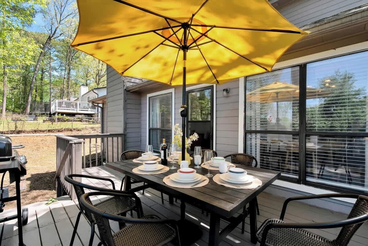 An outdoor dining area with a table set for four, surrounded by wicker chairs, under a yellow umbrella.