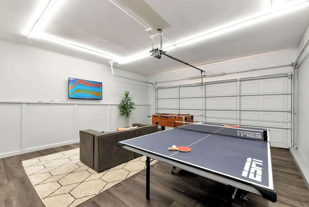 Una mesa de ping pong con una paleta, una mesa de futbolín y un proyector en una habitación con puerta de garaje.