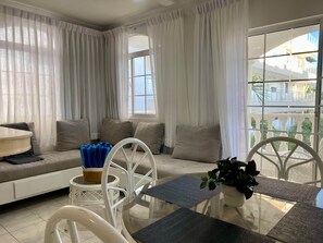 Ein modernes Wohnzimmer mit einer Couch, Sesseln, einem Glastisch und Blick auf ein Gebäude durch große Fenster.