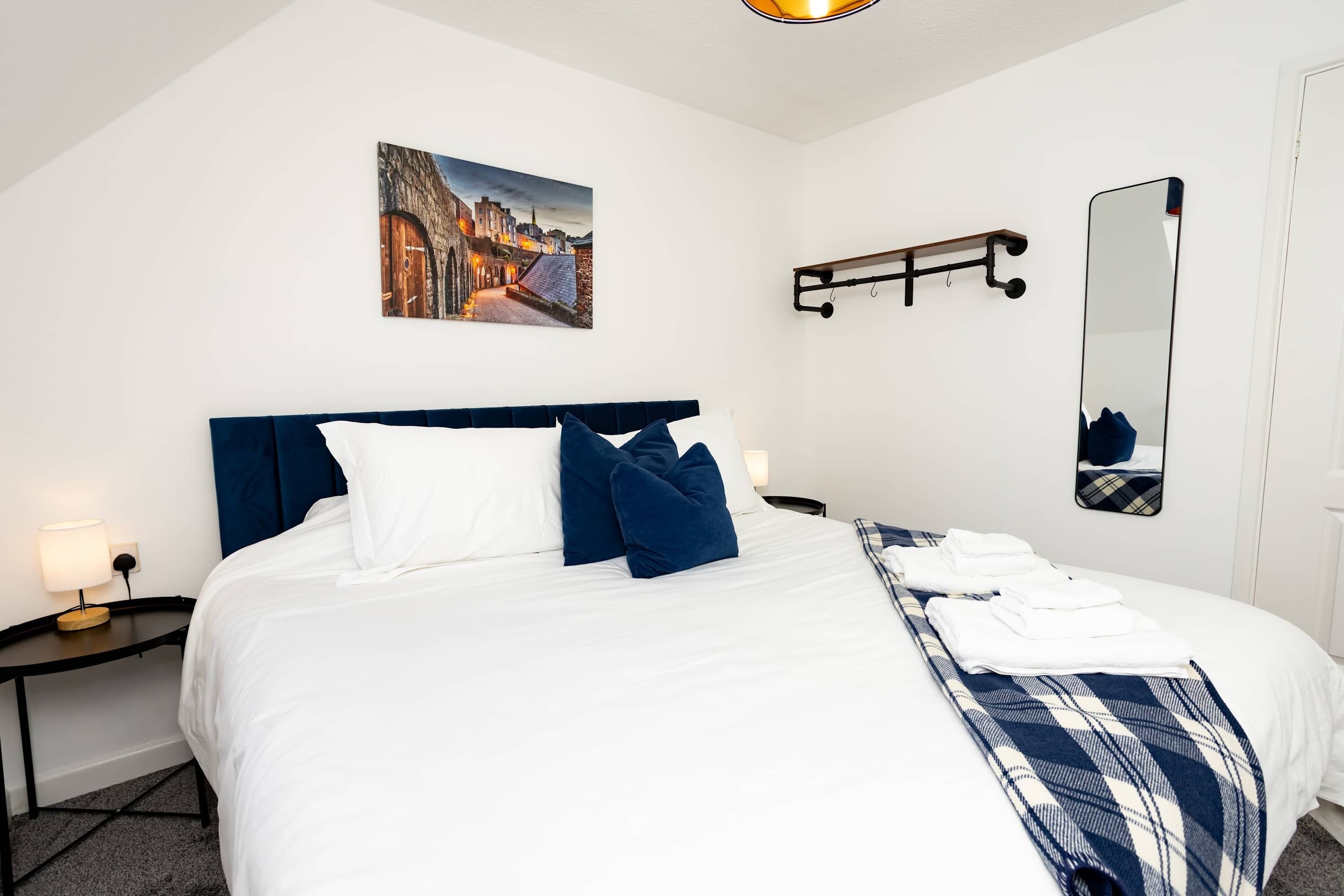 Een hotelkamer met een net opgemaakt bed, een schilderij van een stadsgezicht, een spiegel aan de muur en een klein nachtkastje met een lamp.