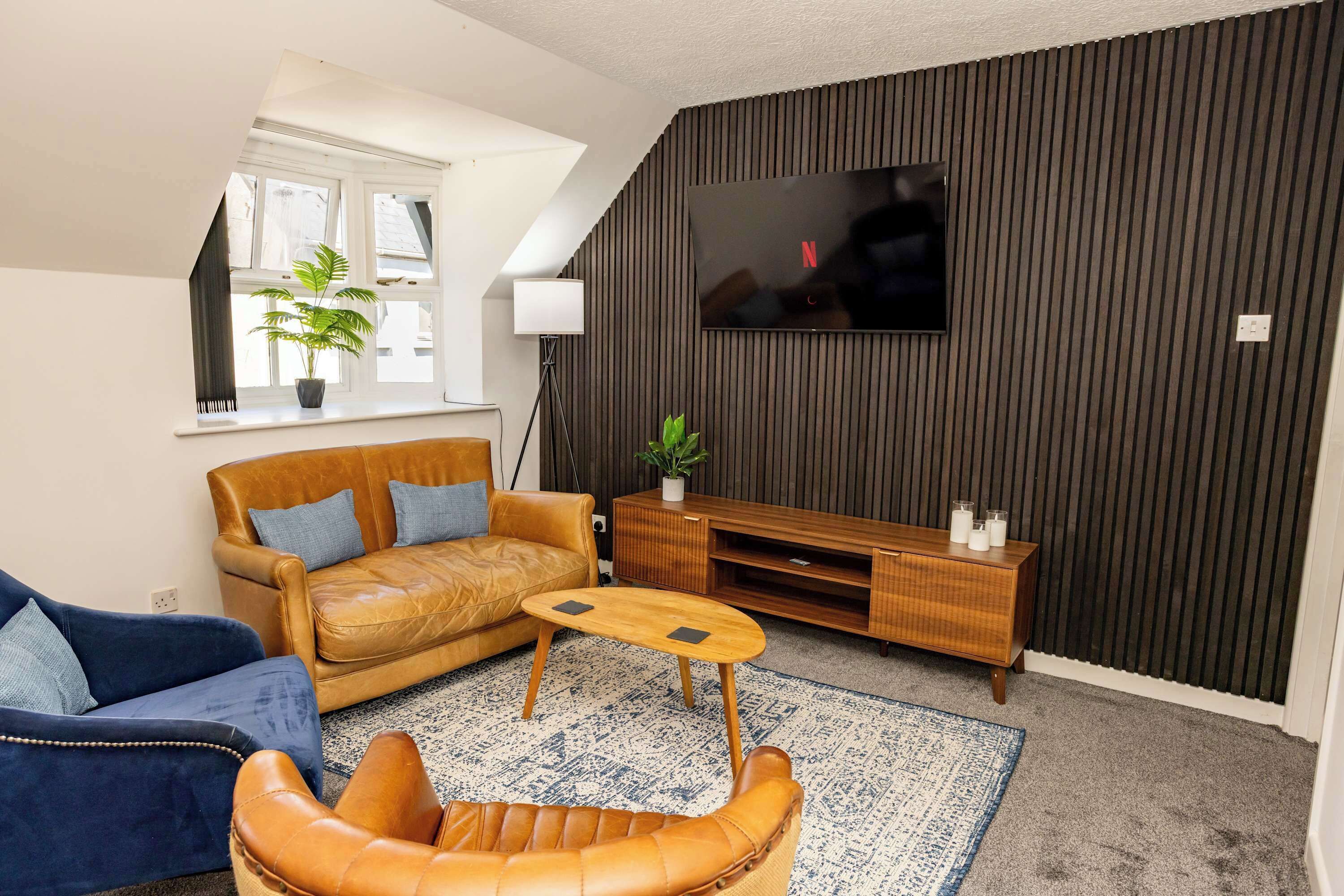 A modern living room with a leather sofa, a blue armchair, a wooden coffee table, a TV mounted on a dark wall, and a potted plant.