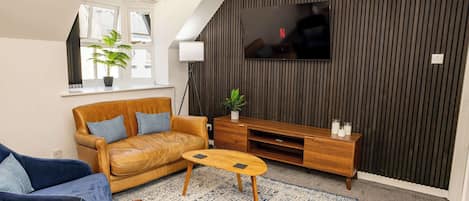 A modern living room with a leather sofa, a blue armchair, a wooden coffee table, a TV mounted on a dark wall, and a potted plant.
