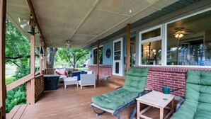 Eine überdachte Veranda mit Holzboden, ein grüner gepolsterter Loungesessel, ein kleiner Tisch und eine Rattan-Couch mit roten Kissen.