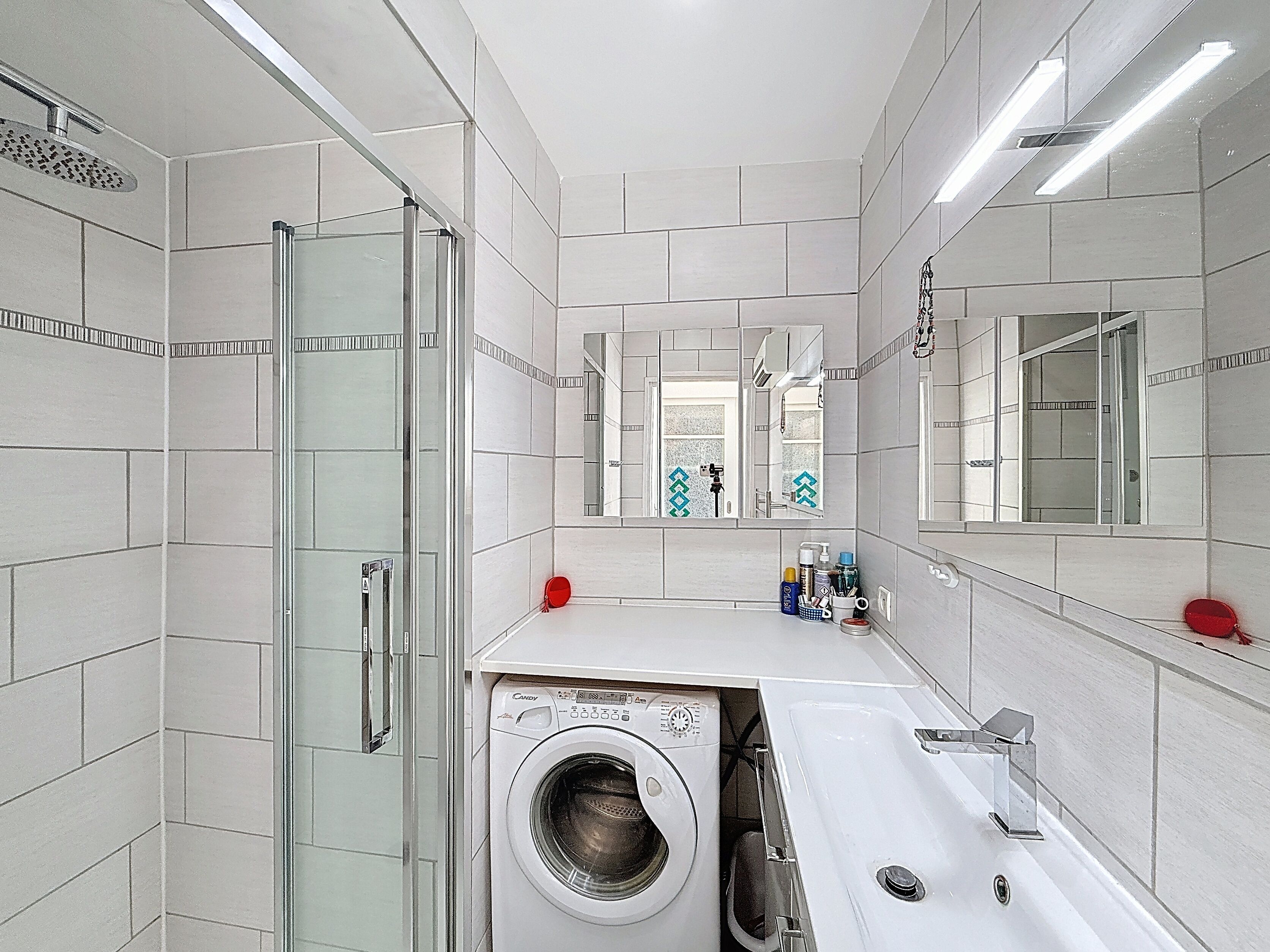 A modern bathroom with a glass shower enclosure, a built-in washing machine, and a large mirror above the sink.