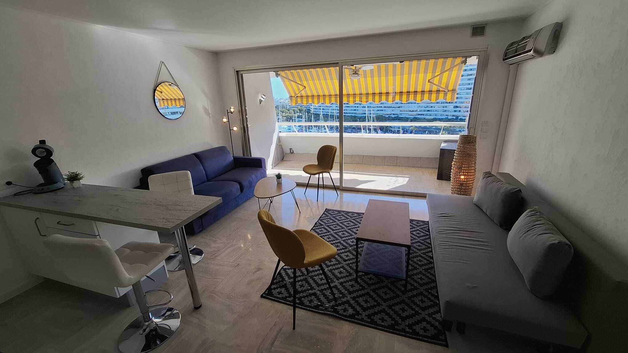 Ein modernes Wohnzimmer mit einer Couch, einem Esstisch und einem Sessel. Ein großes Fenster bietet Blick auf Gebäude und ein gelbes Markisen