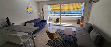 Ein modernes Wohnzimmer mit einer Couch, einem Esstisch und einem Sessel. Ein großes Fenster bietet Blick auf Gebäude und ein gelbes Markisen