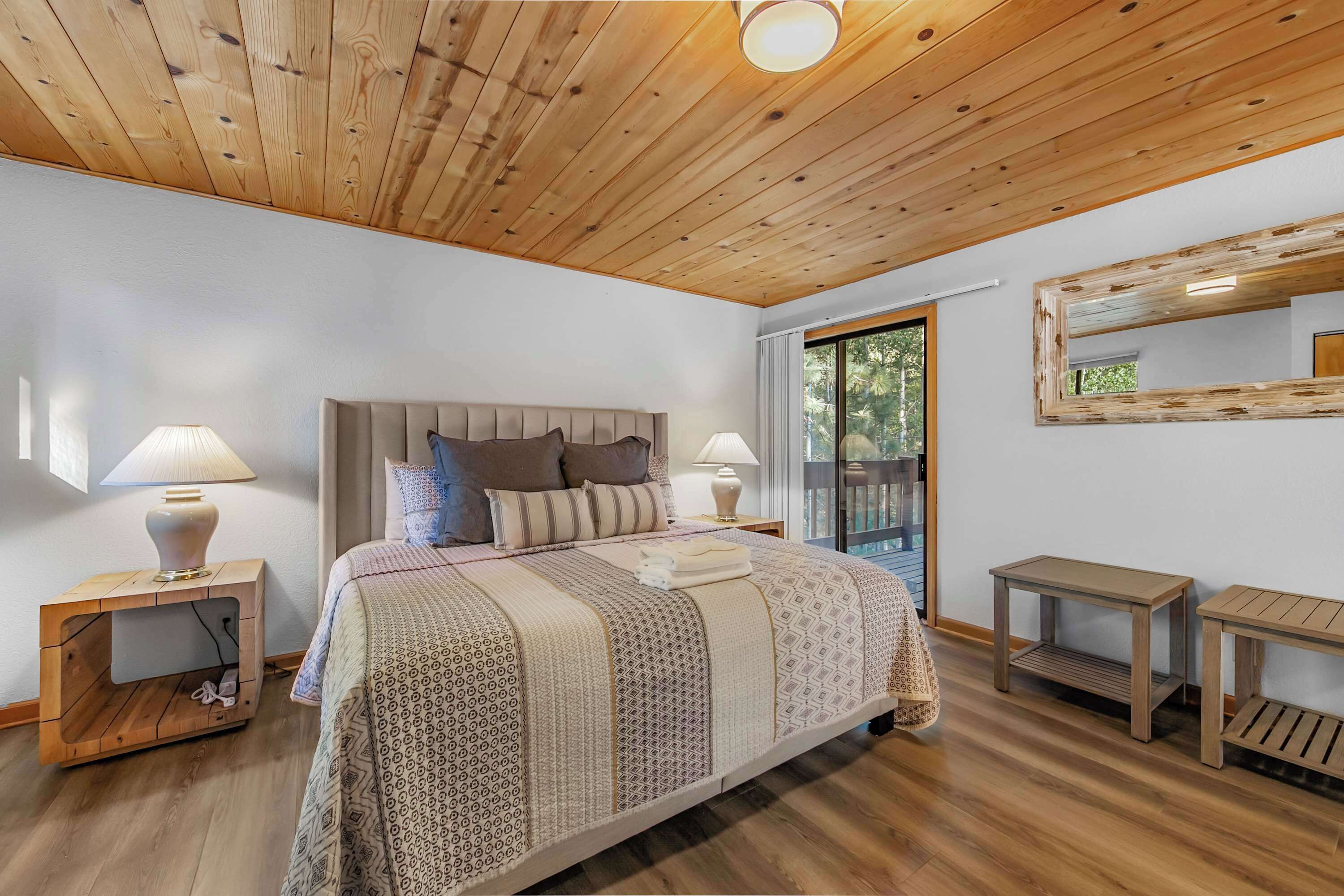 A bedroom with a wooden ceiling, a bed with patterned bedding, two bedside tables with lamps, and a large window.