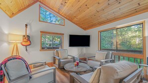 Ein geräumiges Wohnzimmer mit hoher Decke, Holzbalken, einem wandmontierten Fernseher und großen Fenstern mit Blick ins Freie.