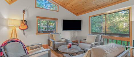 Amplia sala de estar con techo alto, vigas de madera, televisor montado y grandes ventanas con vistas al exterior.