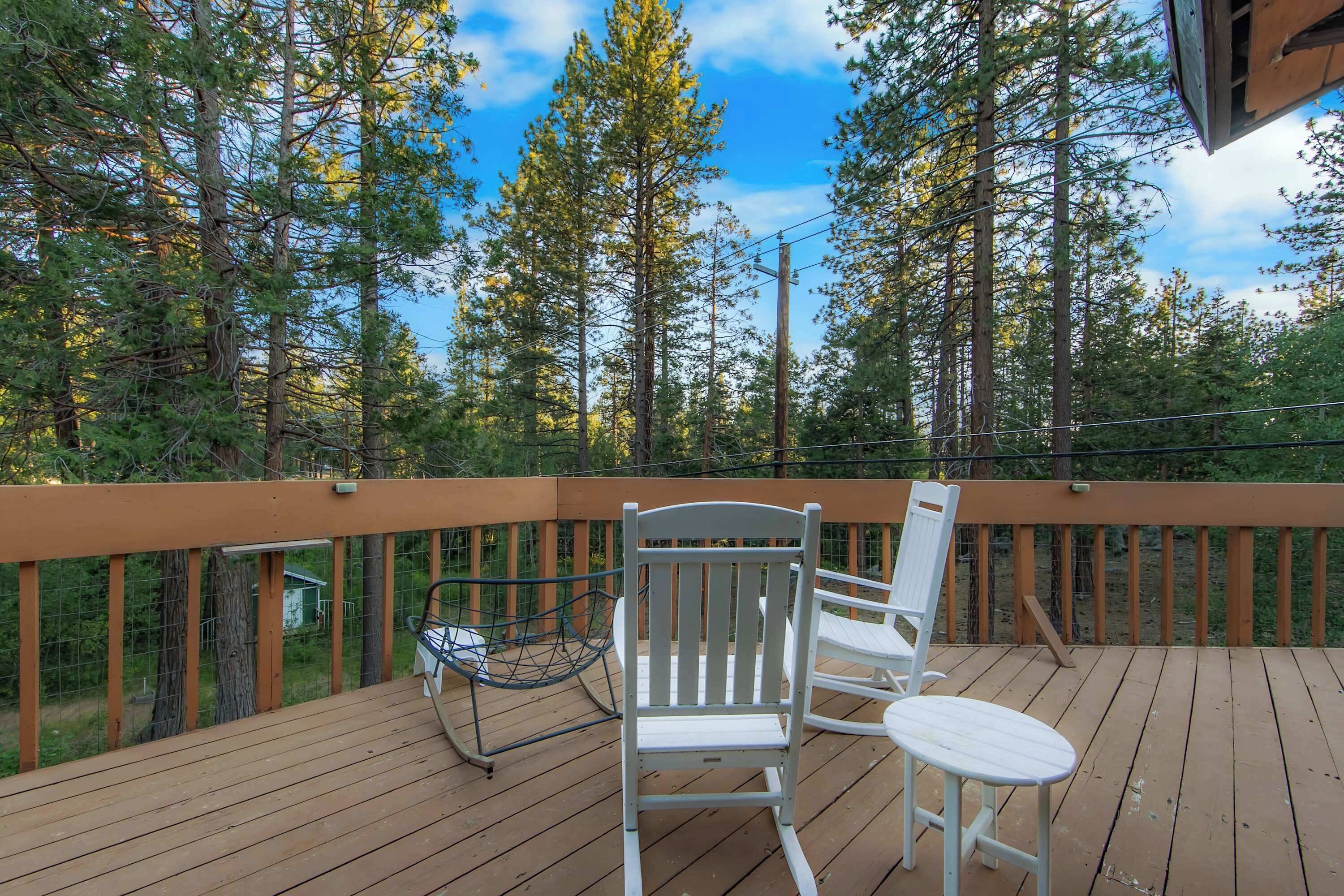 A wooden deck with two white chairs and a small table, surrounded by a wooden railing and overlooking a forest.