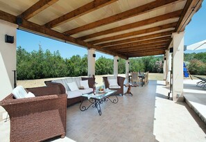 Eine überdachte Terrasse im Freien mit Korbmöbeln, einem Glastisch und Blick auf viel Grün.