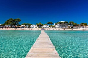 A wooden pier leading to a beach with clear turquoise water, surrounded by buildings and trees. - Impressive & Spacious Villa - Private Pool, Outdoor Kitchen, Bar & Sauna - 20 mins walk to Alcudia! (Alcudia)