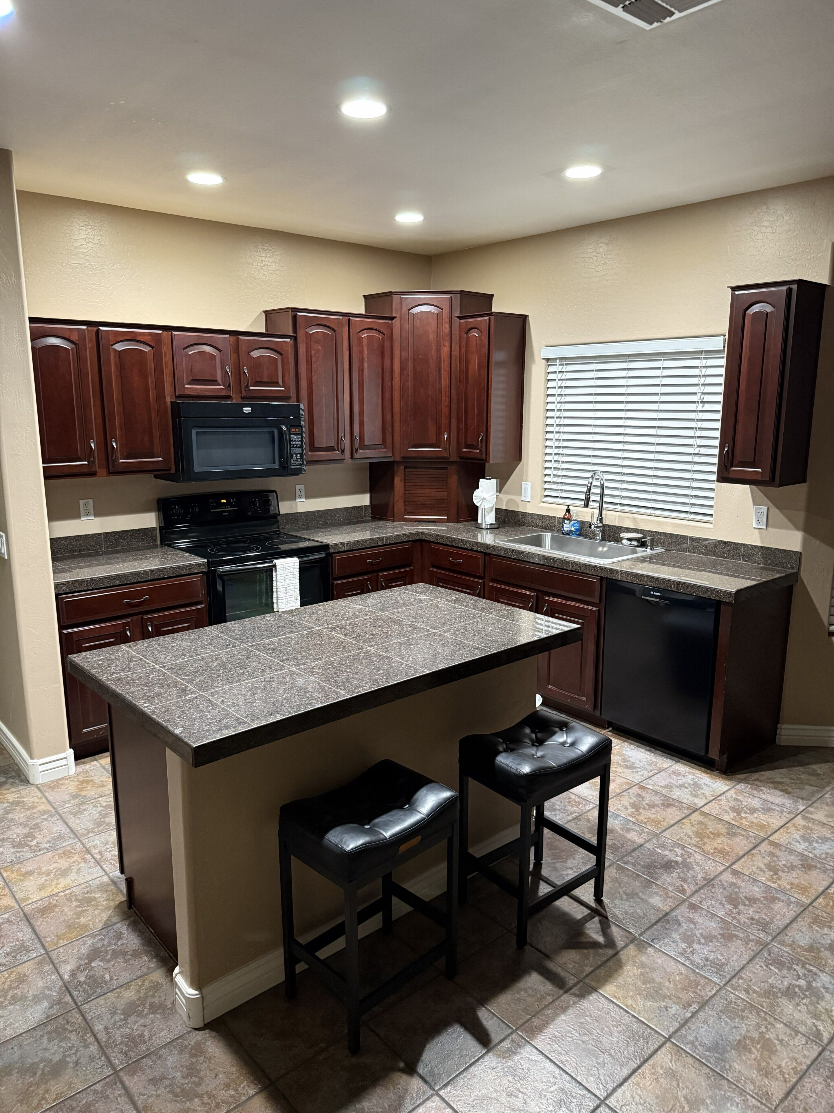 A kitchen with dark wooden cabinets, a granite countertop, a built-in microwave, a dishwasher, and two bar stools.