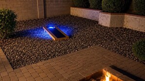 Zona con fogata, fuego encendido, patio de ladrillo y área de grava con una estructura iluminada en azul.