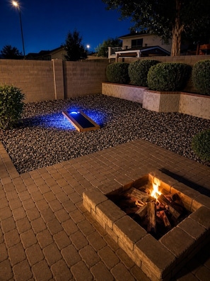 Ein Bereich mit Feuerstelle, in dem ein Feuer brennt, umgeben von einer Pflasterterrasse und einem Kiesbereich mit einer blau beleuchteten Struktur.