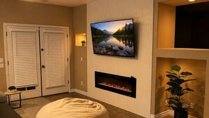 Una sala de estar con una televisión de pantalla plana en la pared, chimenea, un sillón informal y un ventilador de techo.