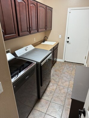 A compact kitchen with a washer and dryer, a sink, and a door. - Peaceful Family Retreat with Hot Tub, Game Room & BBQ Near State Farm Stadium (Avondale)