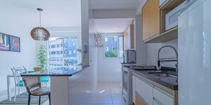 A modern kitchen with a central island, a dining area with a table and chairs, and a view of a building through a window. - Apartamento em Ubatuba com Piscina e Churrasqueira (Ubatuba)