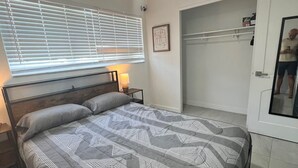 Ein Schlafzimmer mit einem Bett, einem Deckenventilator, einem Fenster mit Jalousien, einer Nachttischlampe und einem Kleiderschrank.
