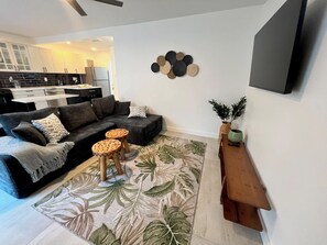 A modern living room with a sofa, two round wooden stools, a coffee table, a patterned rug, a wall-mounted decorative piece, and a small wooden side table with a plant. - Apartment 1505 - Wilton Manors, Ft Lauderdale beaches (Fort Lauderdale)