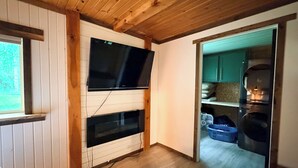 A room with a wooden ceiling, a TV mounted on the wall, a window, and a door leading to a kitchen area. - Lakefront Cabin Near Duluth (Duluth)
