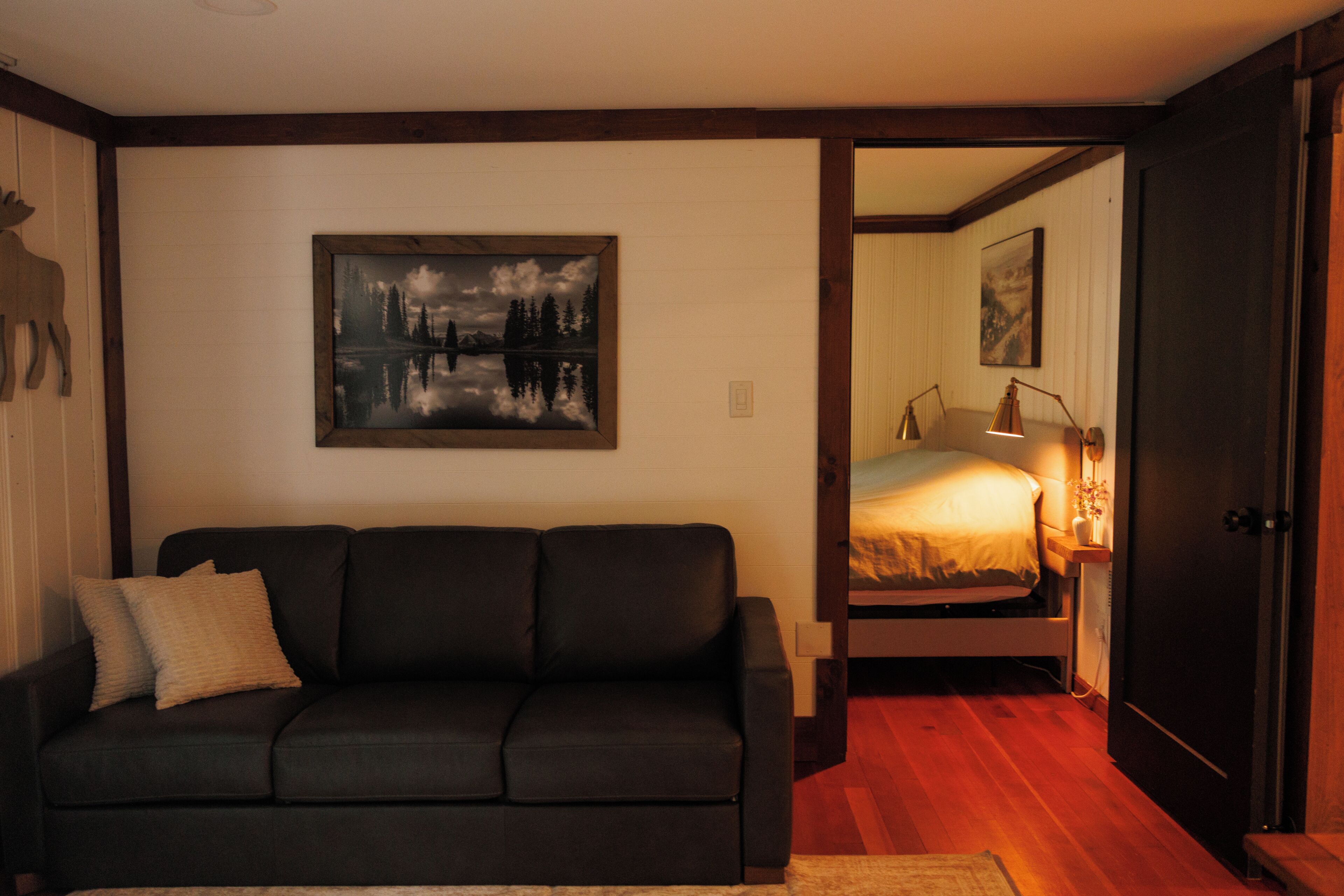 A room with a dark-colored sofa, a painting of a landscape, a bed with a lamp, and a wooden floor.