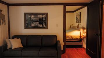 A room with a dark-colored sofa, a painting of a landscape, a bed with a lamp, and a wooden floor.