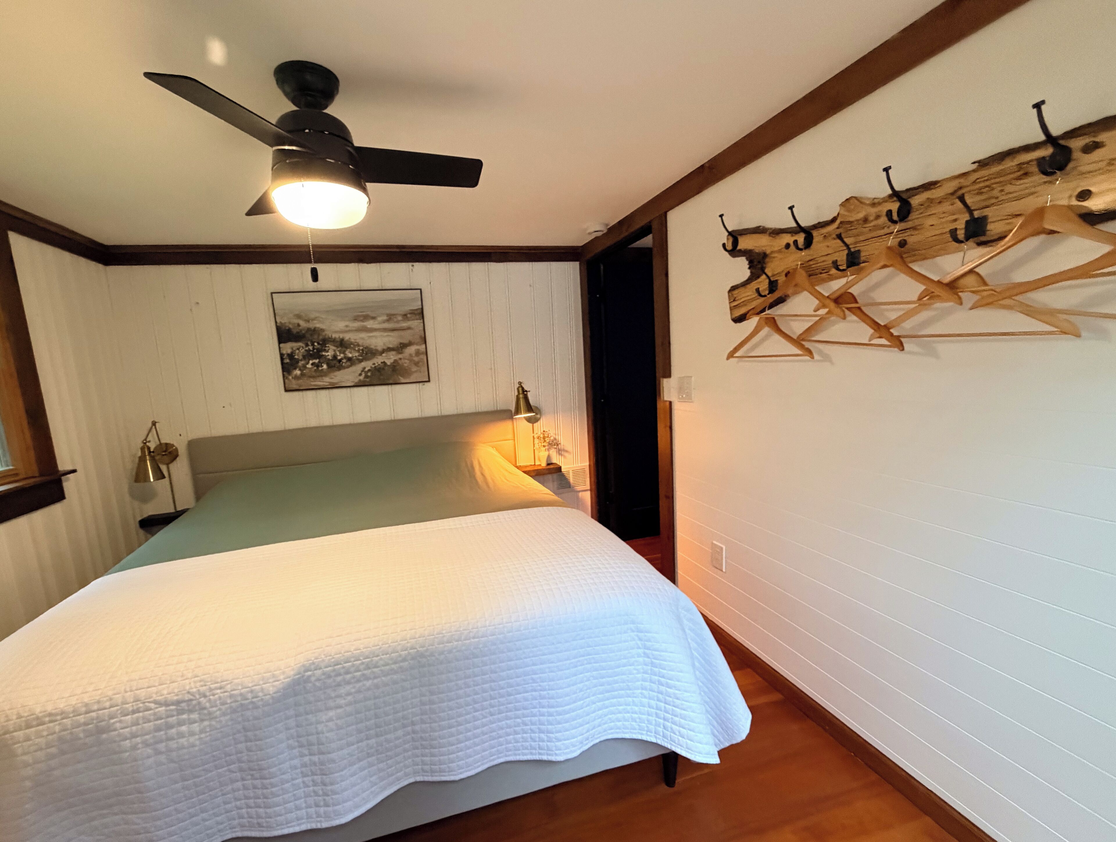 A bedroom with a bed, a ceiling fan, a wall-mounted lamp, a wooden coat rack, and a framed picture.
