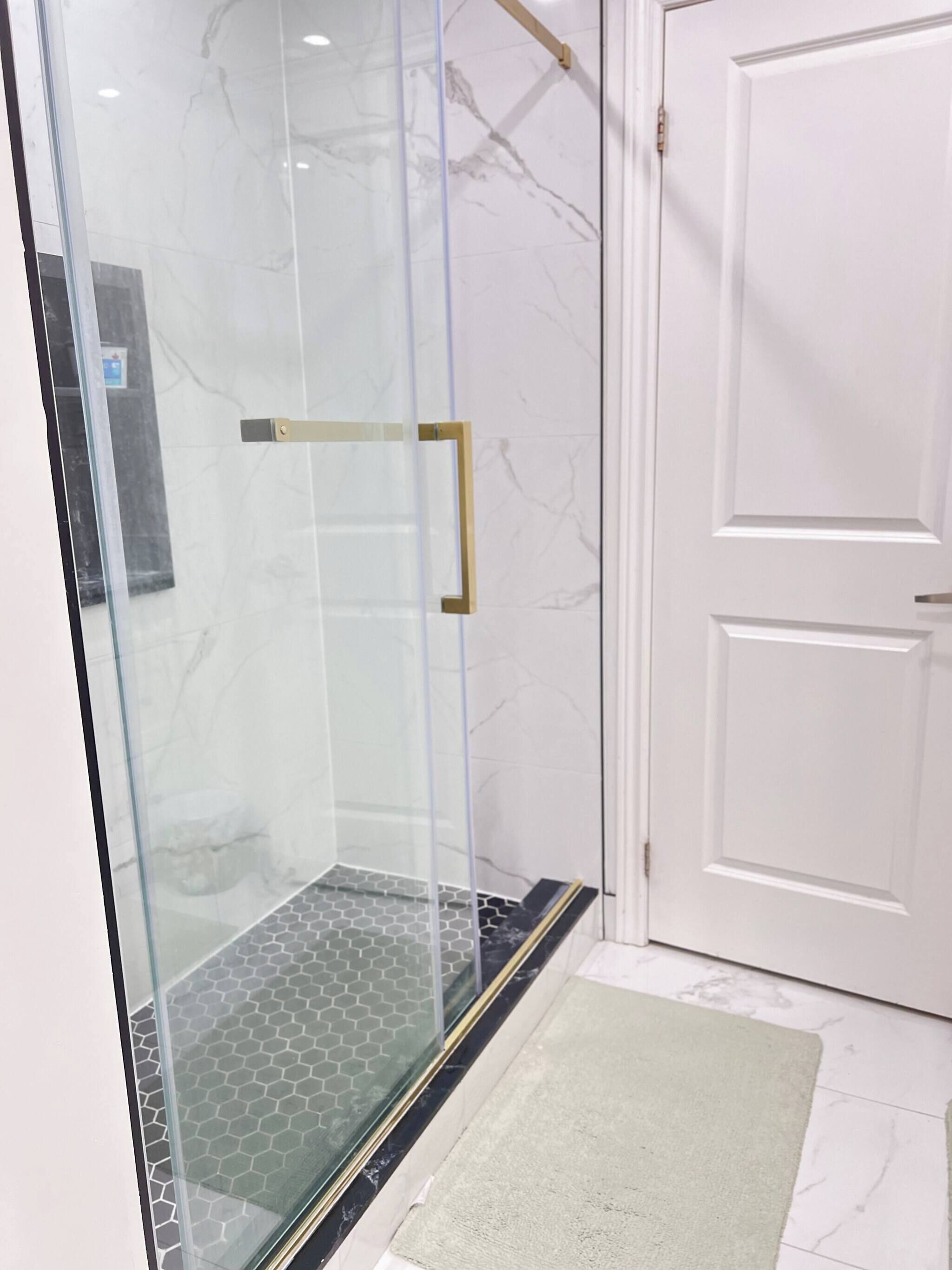 A glass-enclosed shower with a gold handle, a white door with a panel, and a hexagonal patterned floor.