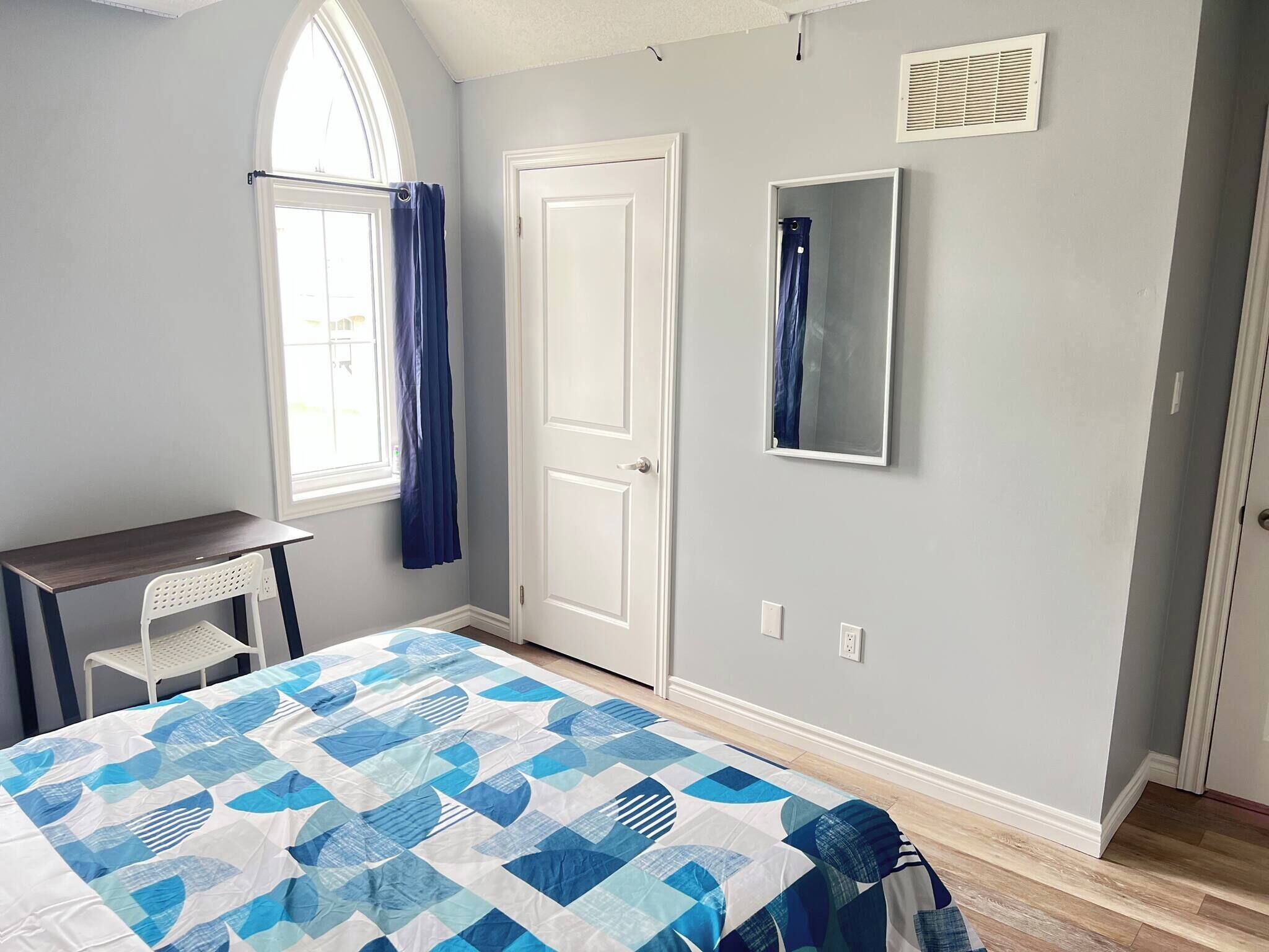 Un dormitorio con cama, escritorio, silla, ventana con cortinas, puerta y espejo en la pared.