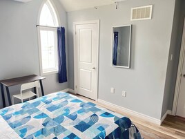 Un dormitorio con cama, escritorio, silla, ventana con cortinas, puerta y espejo en la pared.
