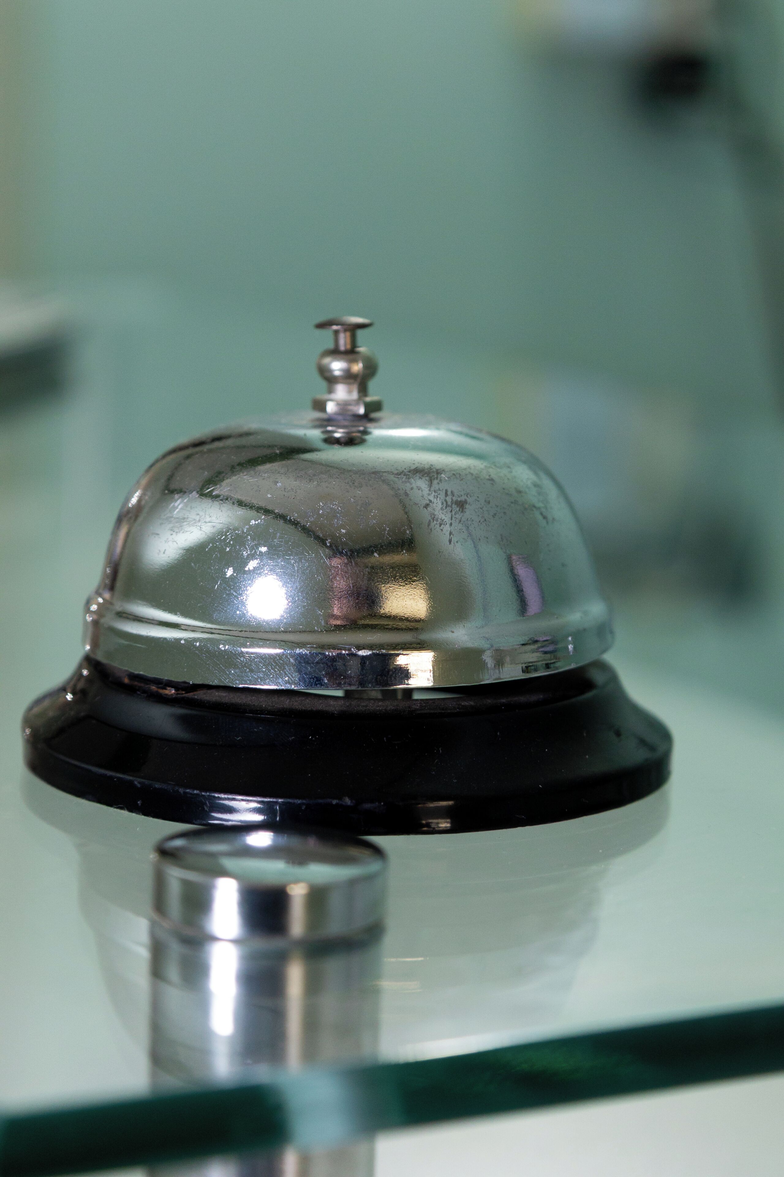 Een metalen bel met een reflecterend oppervlak, gemonteerd op een zwarte voet, waarschijnlijk gebruikt voor het signaleren van roomservice of verzoeken van gasten.