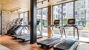 Un gimnasio con cintas de correr, una pared de ladrillo y amplios ventanales con vistas a una zona residencial.