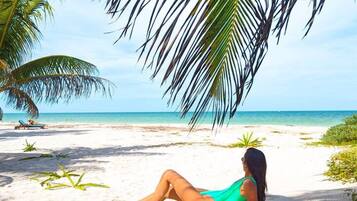 Una persona recostada sobre la arena de una playa, con una palmera encima y un cielo despejado, con un mar tranquilo al fondo.