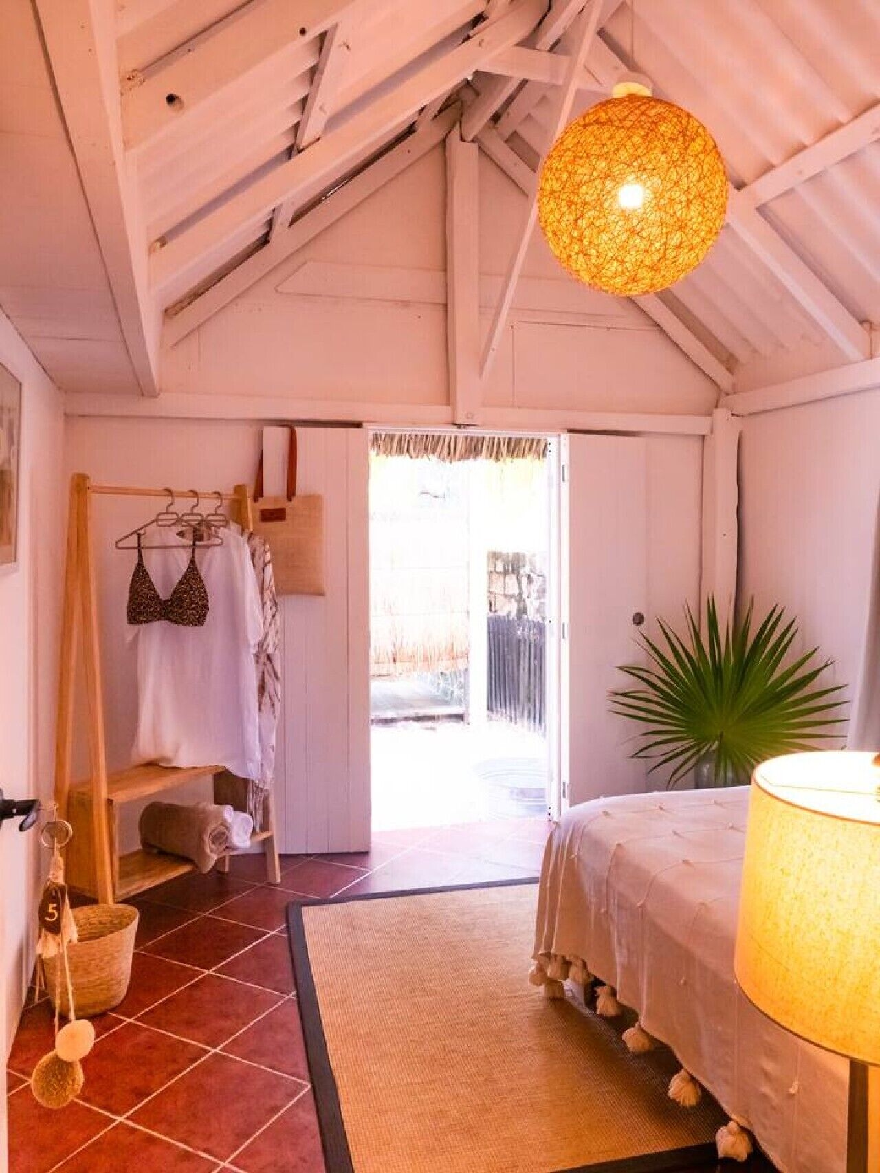 A room with a bed, a hanging light fixture, a wooden rack with clothes, a plant, and a view through an open door.