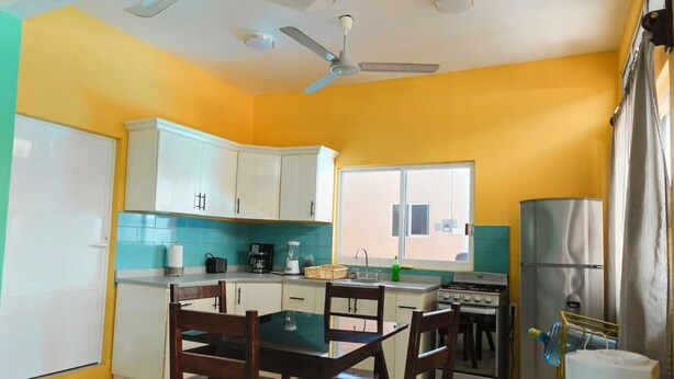 A compact kitchen with white cabinets, a dining table, chairs, a stove, and a refrigerator. - Modern Beachside Retreat • Pool•Parking•Fast Wi-Fi (Playa Agua Blanca)