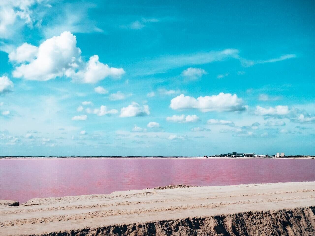 Une vue d’ensemble d’une vaste étendue de sel rose, avec un ciel dégagé et des bâtiments qui se dessinent à l’horizon.