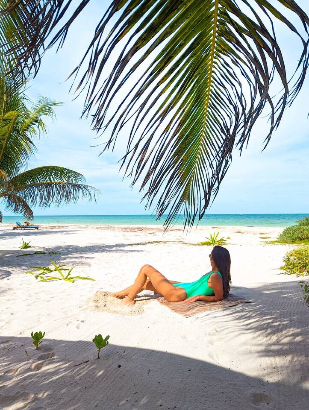 Une personne allongée sur une plage de sable, avec un palmier au-dessus d’elle, un ciel dégagé et une mer calme en arrière-plan.