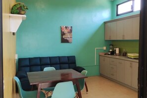 A room with teal walls, a dark blue sofa, a wooden table, and a kitchen area with cabinets and a window.