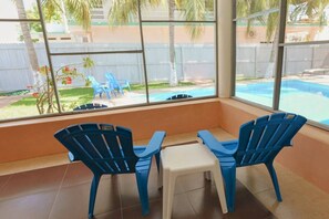 A room with two blue chairs, a small table, and large windows overlooking a swimming pool and a fenced yard.
