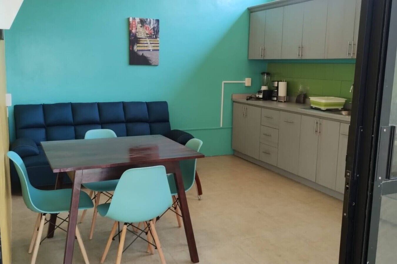 A room with a teal-colored wall, a dark blue sofa, a wooden dining table with four chairs, and a kitchenette area with cabinets and a countertop.