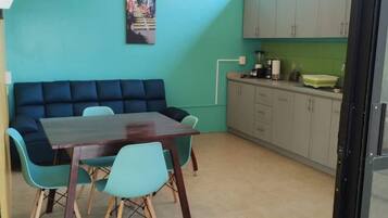 A room with a teal-colored wall, a dark blue sofa, a wooden dining table with four chairs, and a kitchenette area with cabinets and a countertop.