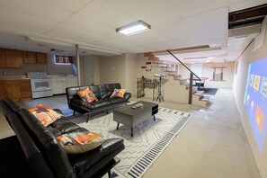 Ein modernes Wohnzimmer mit einer schwarzen Ledersofa, einem Couchtisch und einem gemusterten Teppich. Im Hintergrund ist eine Küche mit Holzschränken und einem Herd erkennbar.