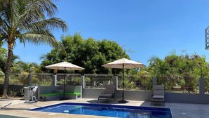 Piscina con sillas de descanso y sombrillas, rodeada por una valla y follaje.