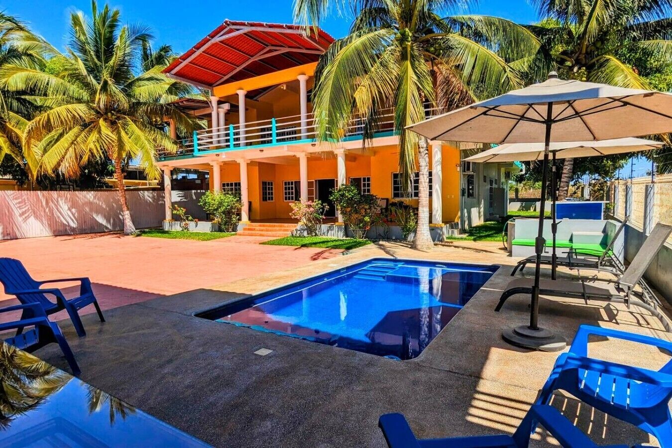 Een zwembadgedeelte met ligbedden en parasols, een gebouw met twee verdiepingen en een rood dak, en palmbomen op de achtergrond.