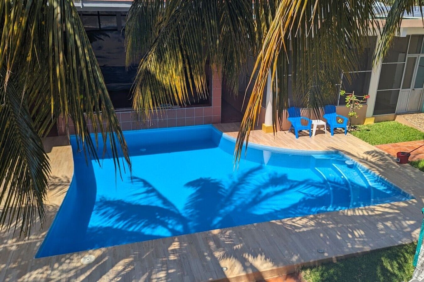 Et rektangulært swimmingpool med en træterrasse hele vejen rundt, palmer og et lille terrassemiljø med stole.