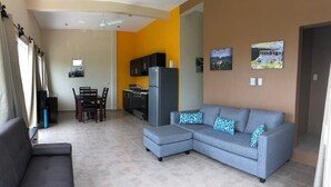 A living room with a grey sectional sofa, a dining area with a table and chairs, a kitchen with black cabinets and a fridge, and artwork on the walls. - Spacious 2BR/2.5BA Oceanview Casita with Terrace (Playa Agua Blanca)
