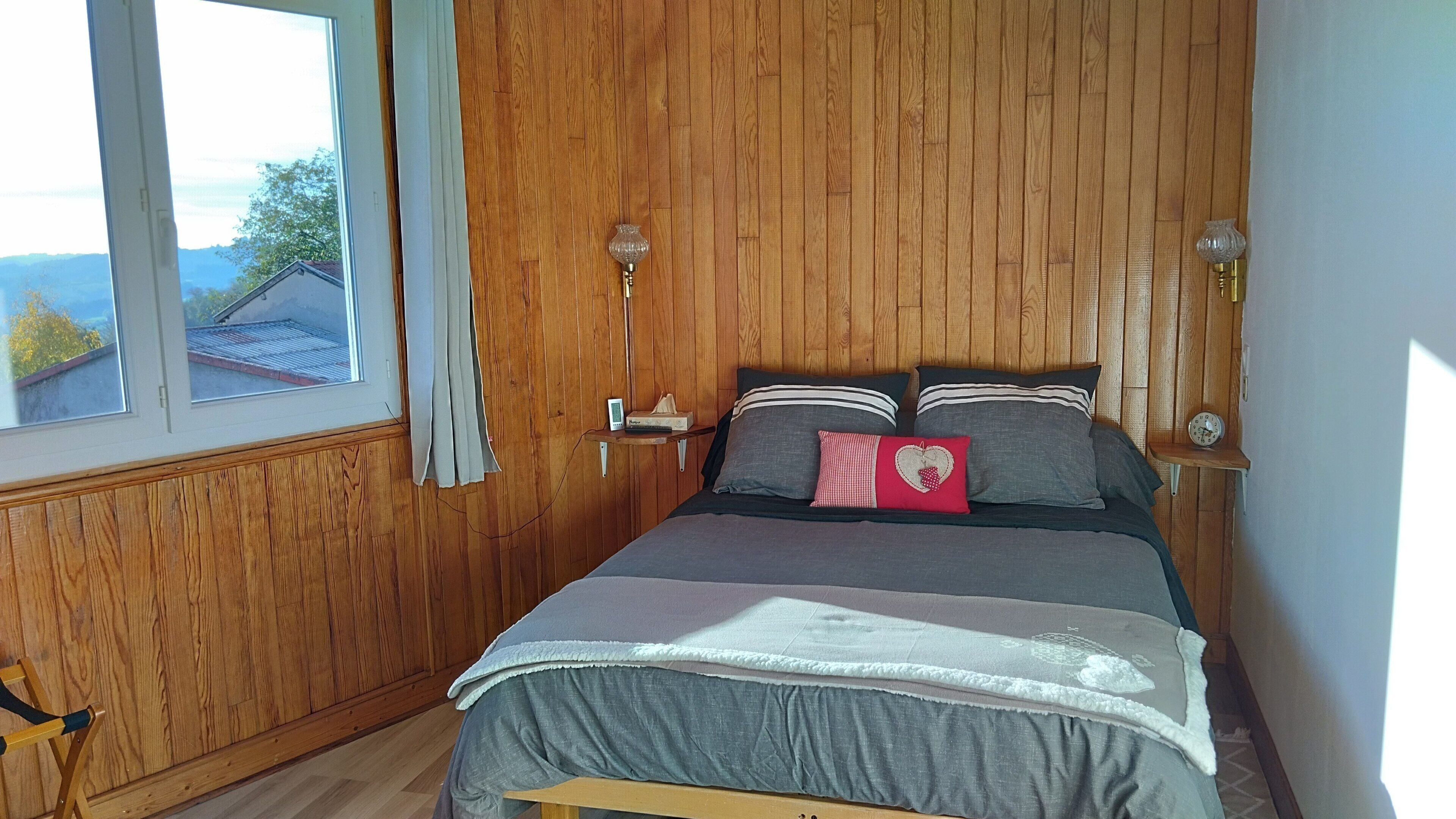 Ein Schlafzimmer mit Holzwänden, einem Bett mit grauem Bettzeug, zwei Nachttischlampen und einem Fenster mit Vorhängen.