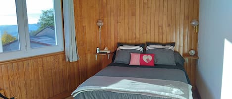 Ein Schlafzimmer mit Holzwänden, einem Bett mit grauem Bettzeug, zwei Nachttischlampen und einem Fenster mit Vorhängen.