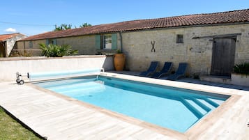A rectangular swimming pool with a wooden deck, a stone building with a tiled roof, and a large planter.